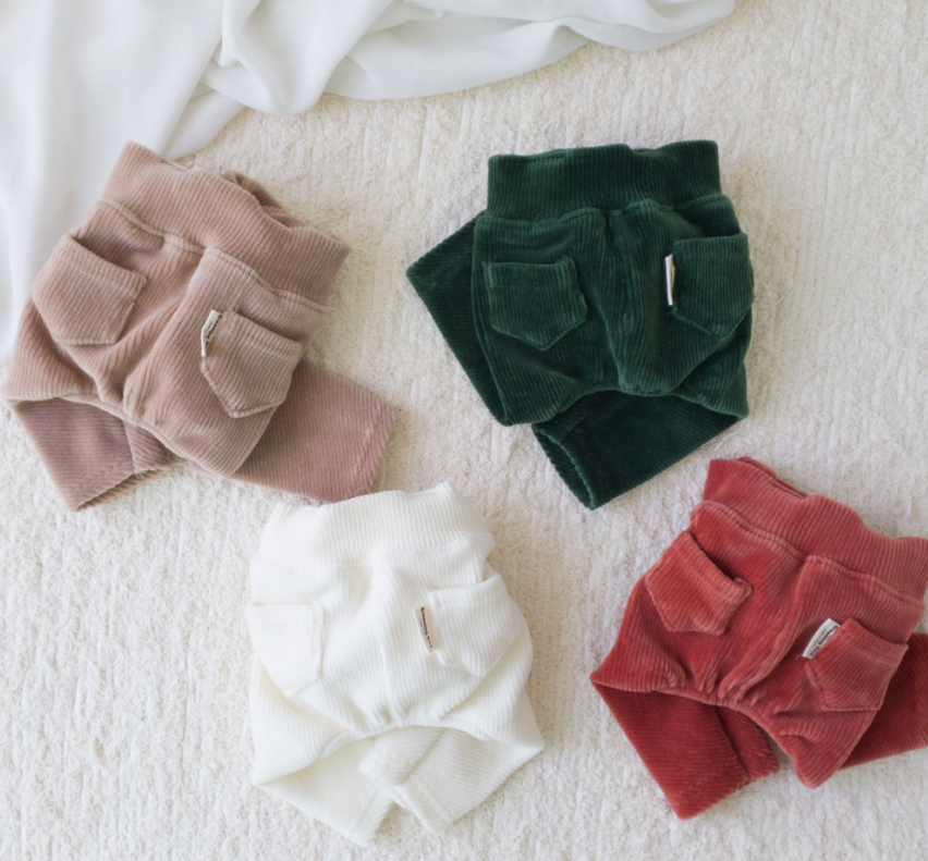 Three pairs of NKpup corduroy dog suspender overalls in mauve, forest green, and terracotta red.
