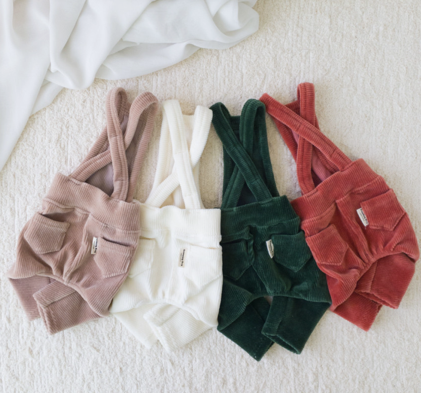 Four NKpup corduroy dog suspender overalls in beige, cream, dark green, and rust red.