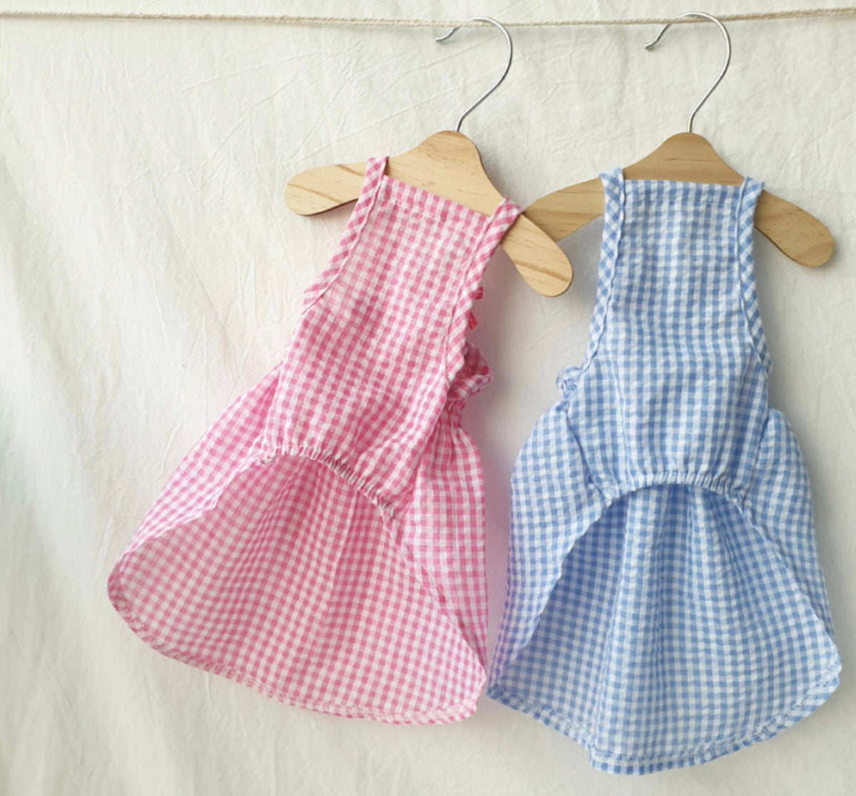 Two vintage gingham dog dresses in pink and blue hanging on wooden hangers.