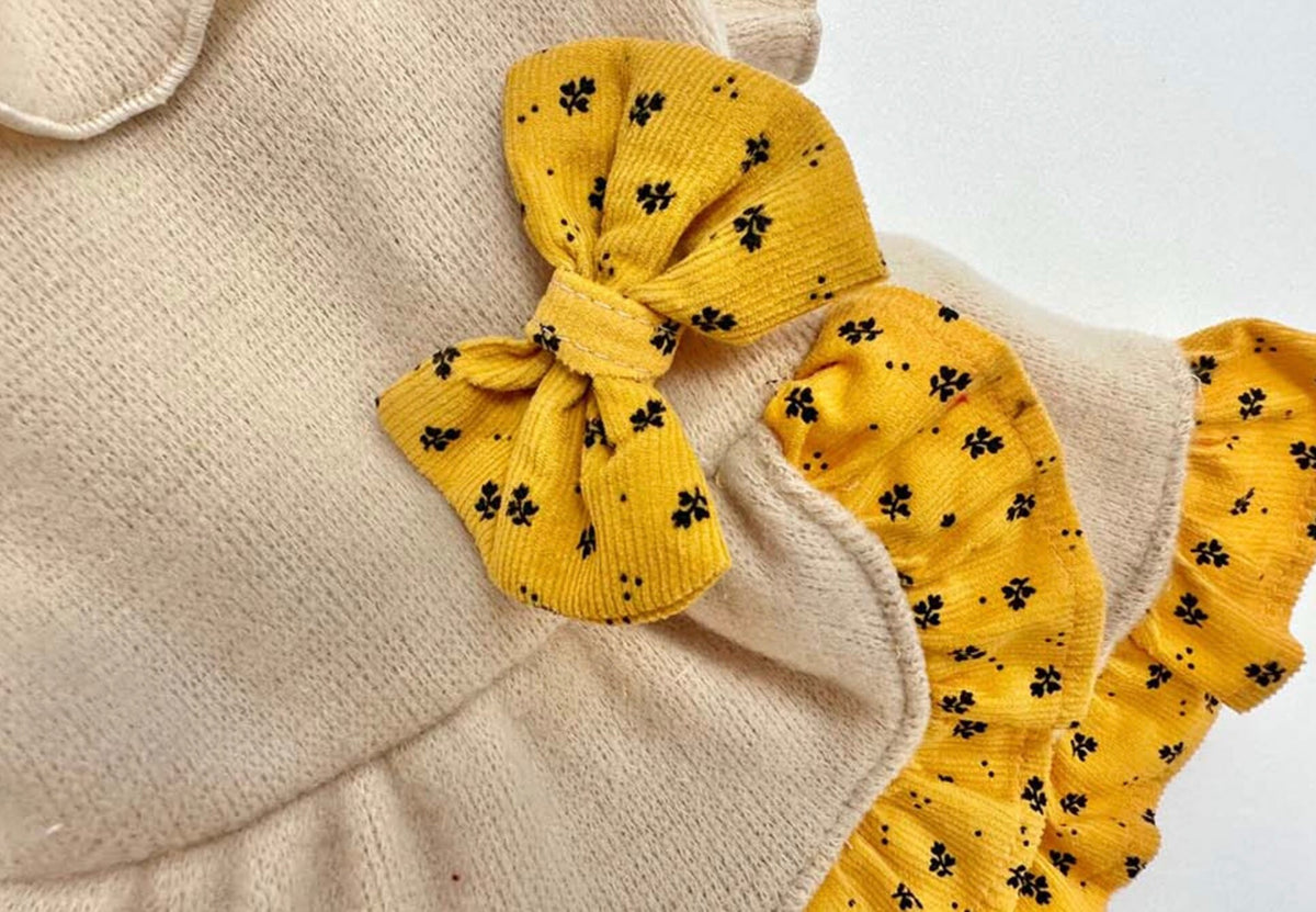 Close-up of a cream NKpup frilled ribbon dog dress with yellow floral patterned bow and trim.