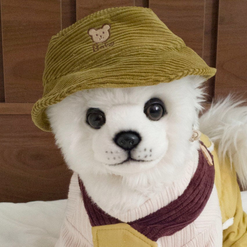 A white dog wearing a vintage mustard yellow corduroy bucket hat with a small bear embroidery.