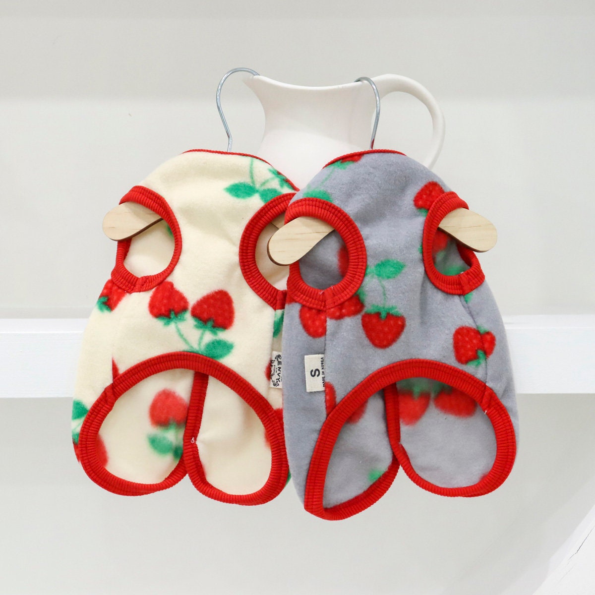 Two strawberry patterned boa fleece dog vests in cream and grey with red trim.