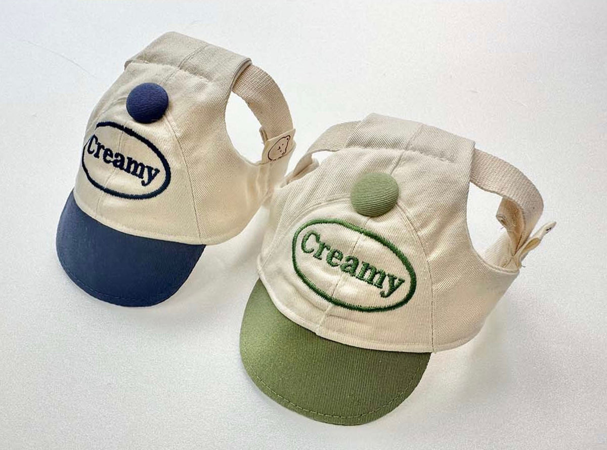 Two Creamy dog cap hats, one in navy blue and one in sage green, featuring embroidered text and a button detail.