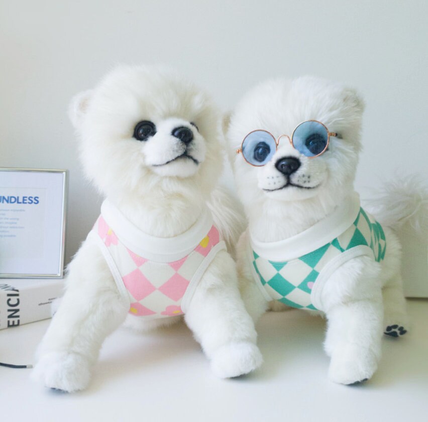 Two white dogs wearing NKpup checkerboard pattern dog shirts in pink and teal colors.