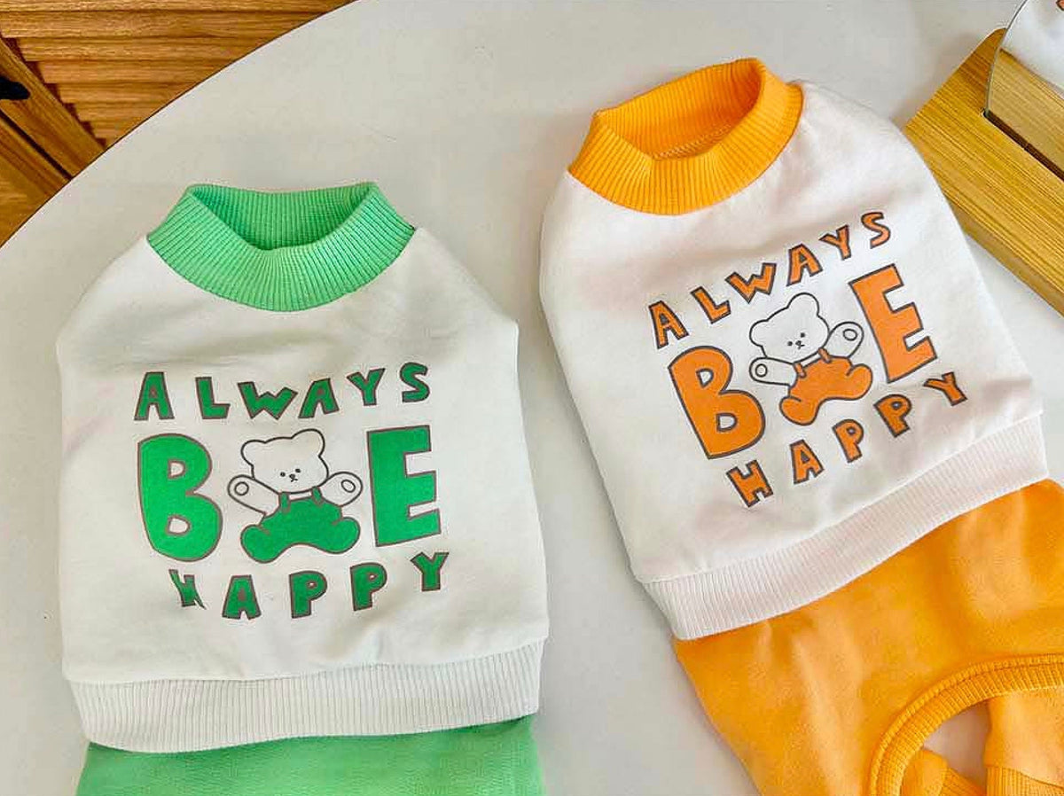 White Always Be Happy dog overalls with green and orange ribbed trim and graphic teddy bear print.