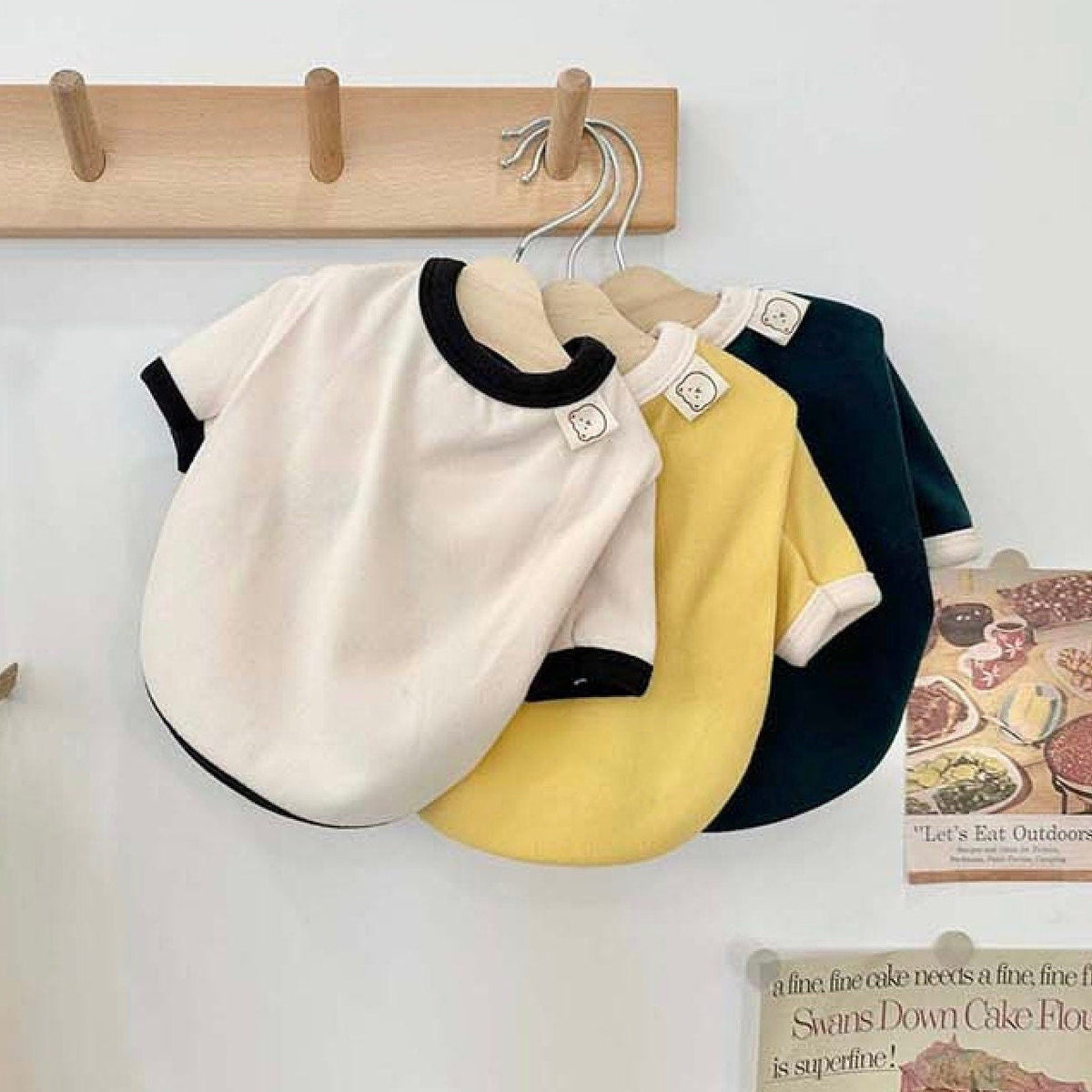 Three NKpup Basic Contrast Tee dog shirts in white, yellow, and dark green hanging on a wooden rack.