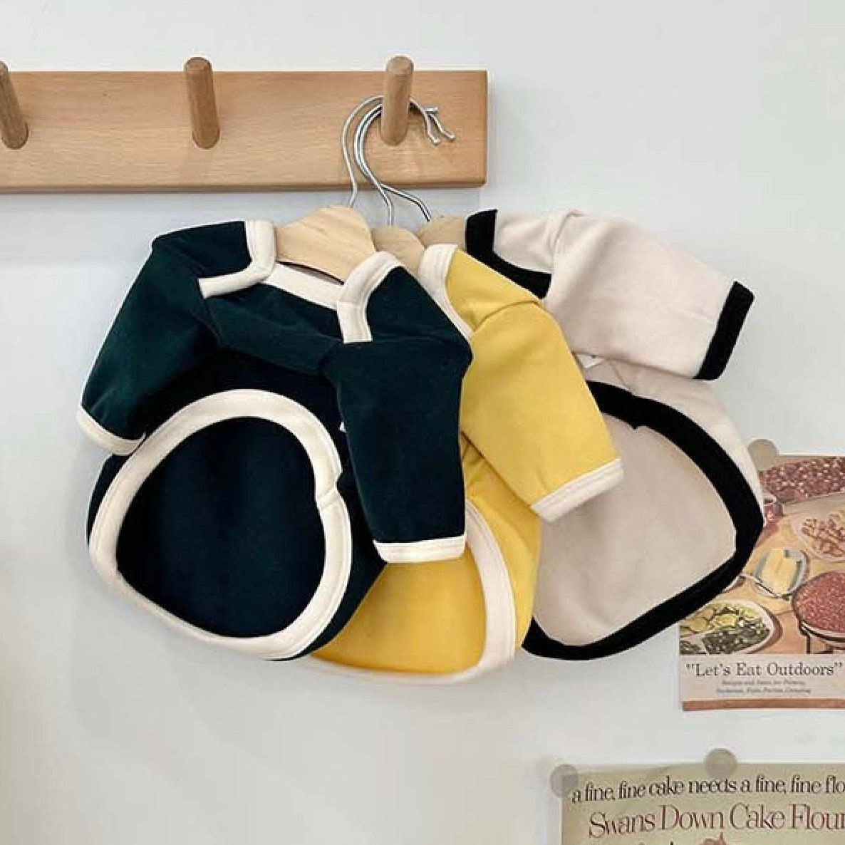 Three NKpup Basic Contrast Tee dog shirts in dark green, yellow, and cream hanging on a wooden rack.