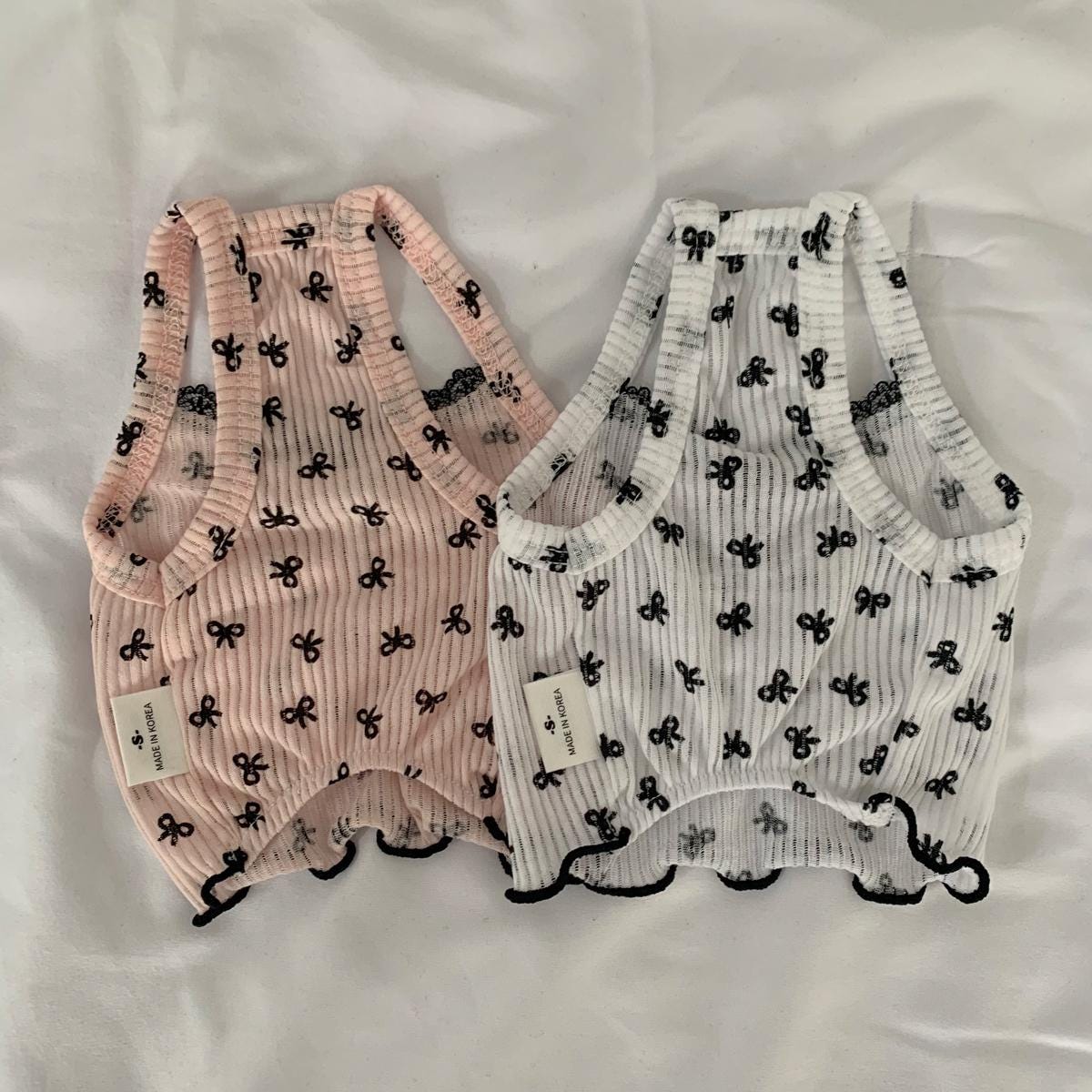 Two NKpup bow crop top dog shirts, one in light pink and one in white, featuring a black bow pattern.