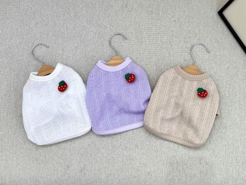 Three NKpup strawberry crop dog cardigans in white, purple, and beige hanging on hangers.