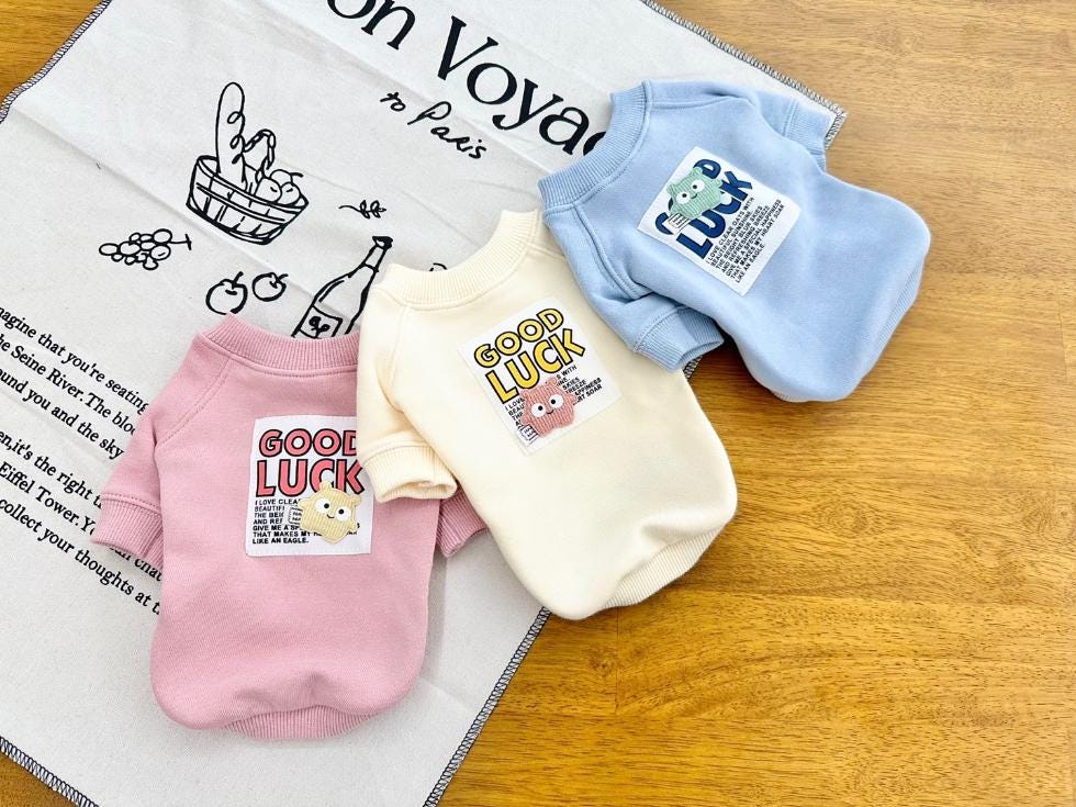 Three NKpup Good Luck sweatshirts for dogs in pink, cream, and light blue.
