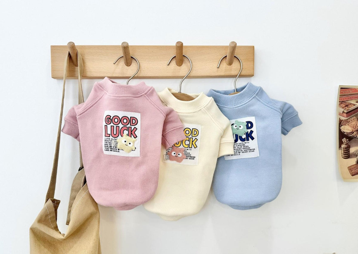 Three NKpup Good Luck sweatshirts for dogs in pink, cream, and light blue hanging on a wooden rack.