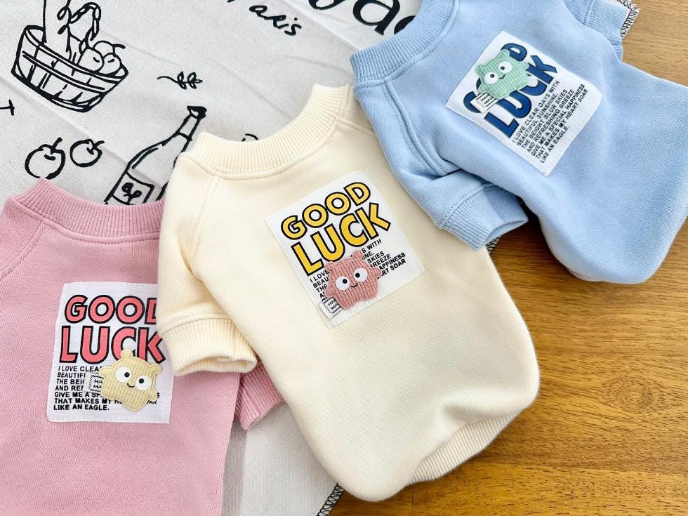 Three NKpup Good Luck sweatshirts for pets in pink, cream, and light blue.