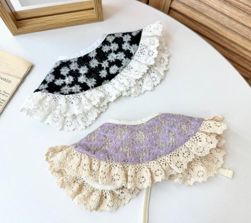 Two floral lace dog capes, one in black with white flowers and one in lavender with purple flowers.