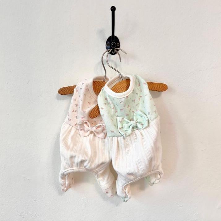 May include: Two pet rompers hanging on a wall hook. One is light pink with a floral pattern and a bow, the other is light green with a floral pattern and a bow. Both rompers are white with a ribbed texture.