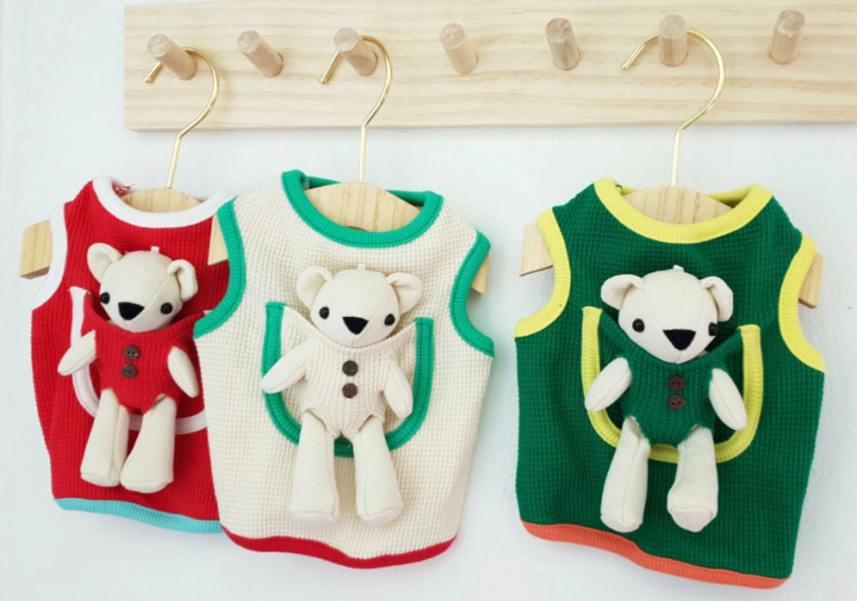 Three NKpup plush teddy bear dog tank tops in red, cream, and green hanging on wooden hangers.