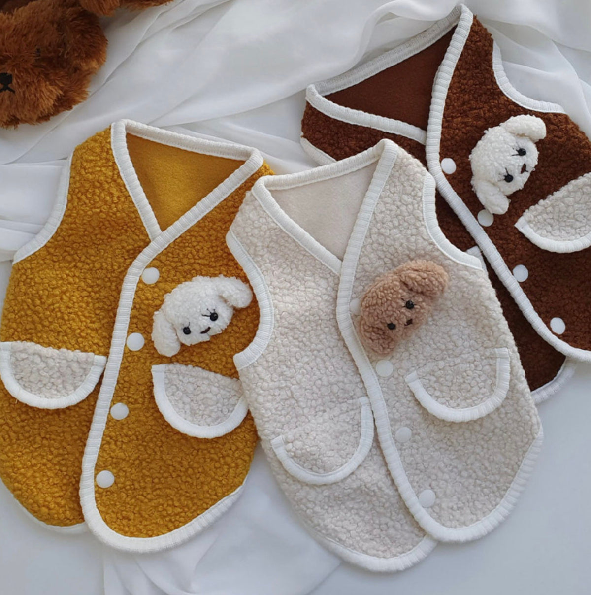 Three plush fleece dog vests in mustard yellow, cream, and brown with adorable bear appliques.