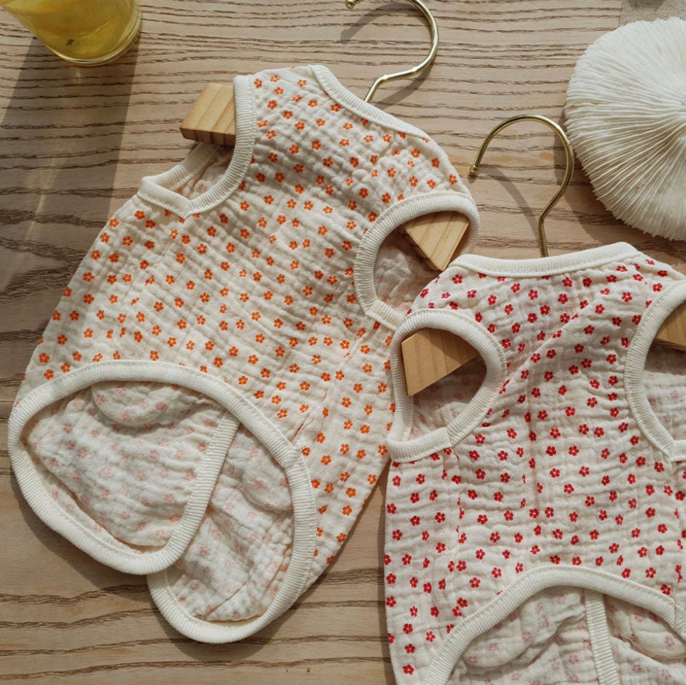 Two crinkle floral dog vests in white with orange and red flower patterns hanging on wooden hangers.