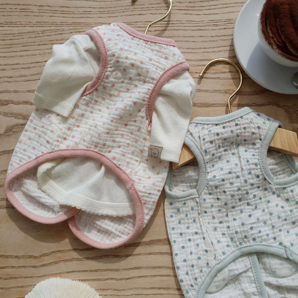 Two polka dot crinkle dog vests in pink and light blue hanging on gold hangers.