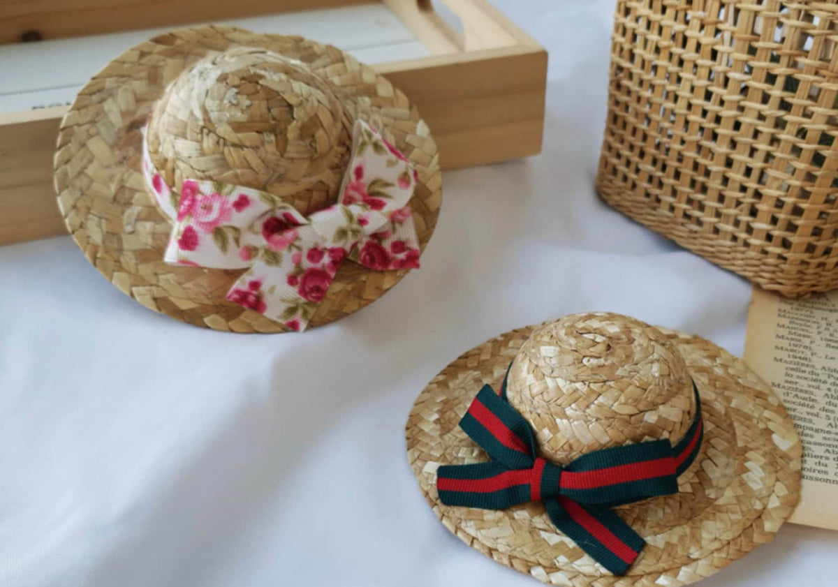 Two straw ribbon dog hats, one with a pink floral bow and one with a green and red striped bow.