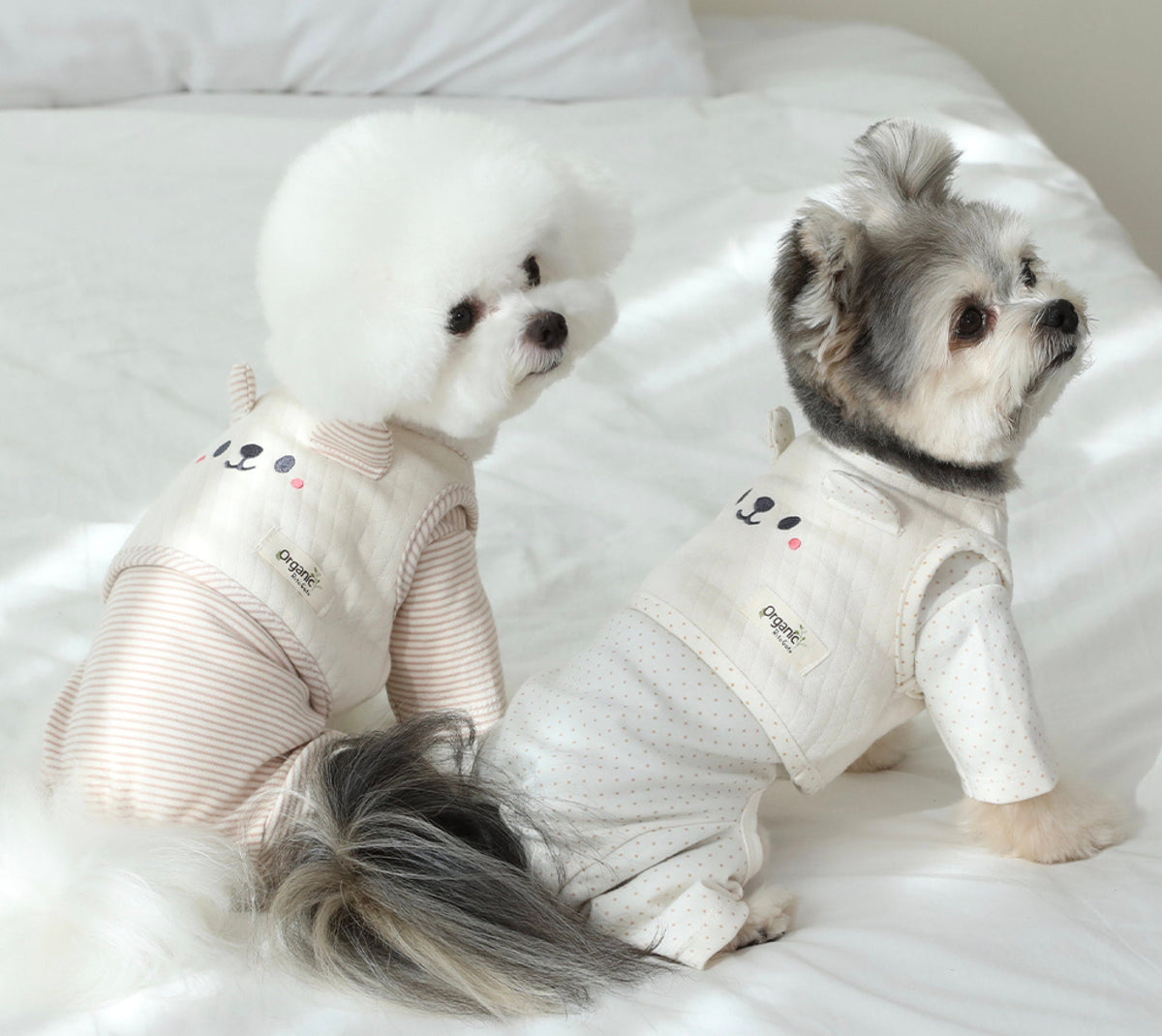 Two dogs wearing NKpup organic quilted dog crop vests in white and light pink striped patterns.